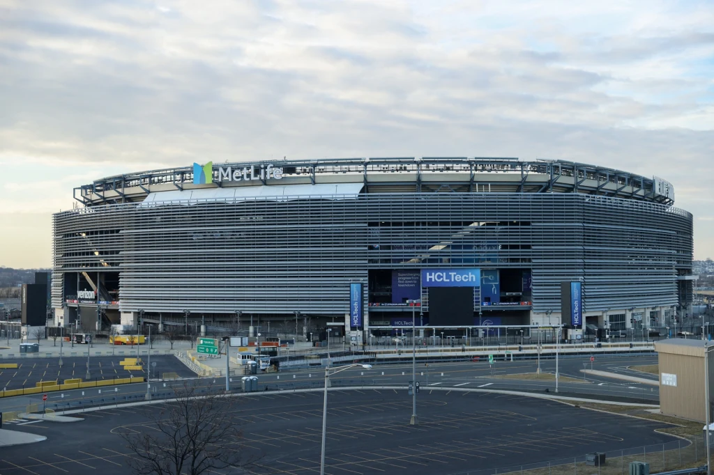MetLife Stadium. To na tym obiekcie zostanie rozegrany finał mistrzostw świata 2026 Nowoczesny stadion o stalowej fasadzie z widocznymi logotypami sponsorów, otoczony pustymi parkingami i wyraźnie wyznaczonymi drogami, pod zachmurzonym niebem.