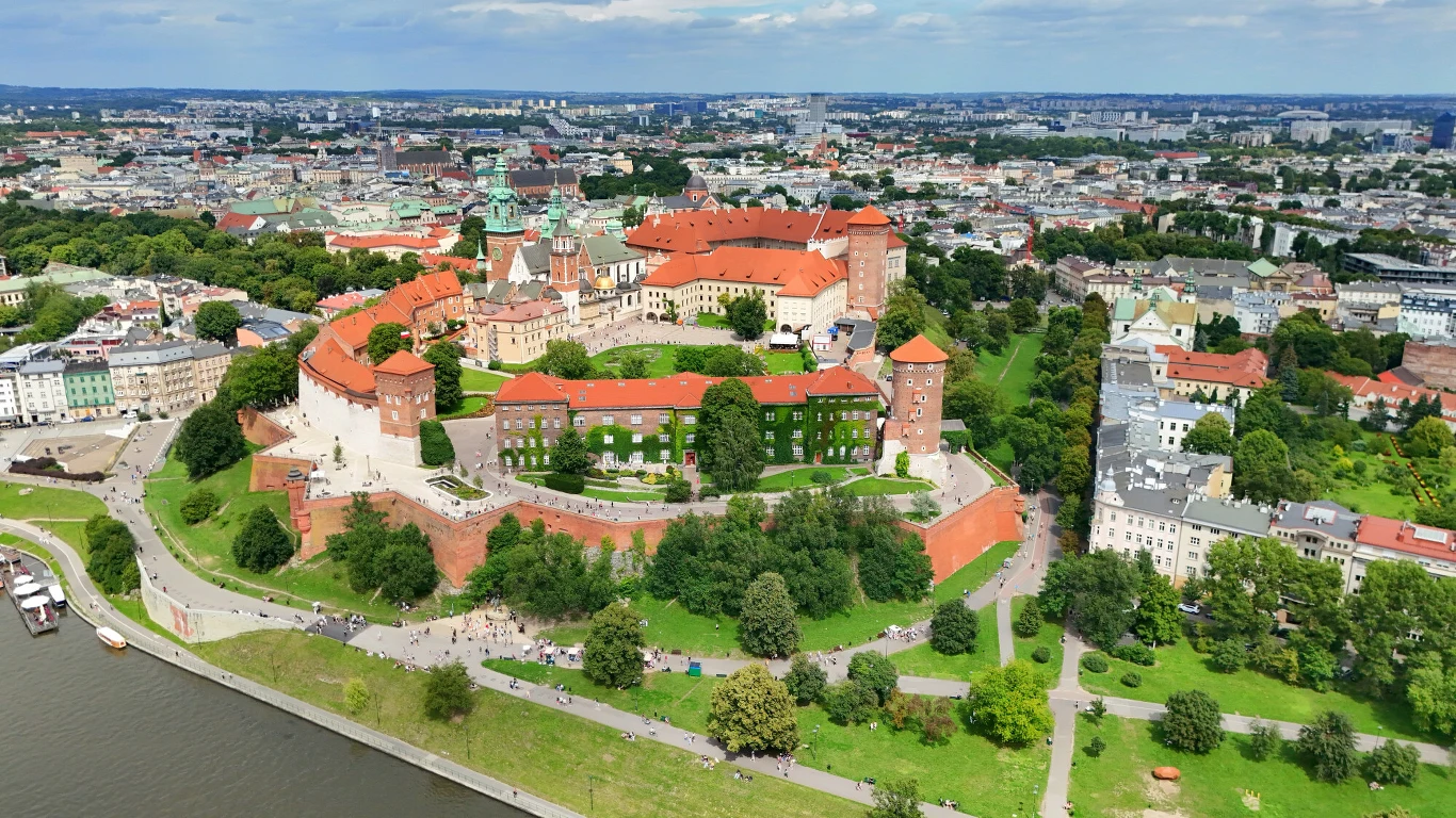 Zamek królewski na Wawelu otoczony murami, z czerwonymi dachami budynków i zielonymi ogrodami, po lewej stronie widoczna Wisła i spacerujące osoby, wokół historycznego obiektu rozpościera się panorama miejskiej zabudowy Krakowa.
