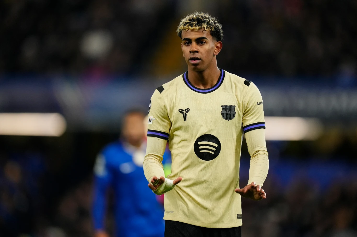 Piłkarz w jasnej koszulce z herbem FC Barcelony i logo sponsora na piersi stoi na murawie podczas meczu piłkarskiego, w tle rozmyty zawodnik drużyny przeciwnej.