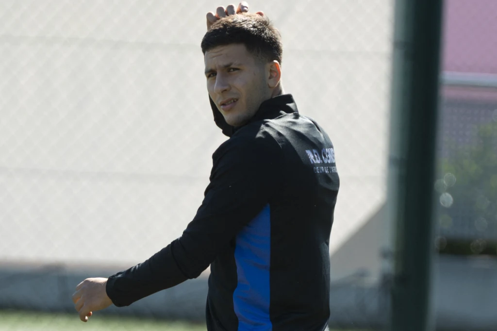 Mężczyzna ubrany w ciemną kurtkę sportową z napisem na plecach stoi na boisku, patrzy przez ramię na fotografa i lekko unosi jedną rękę. W tle widoczna siatka ogrodzeniowa i rozmazane elementy infrastruktury sportowej.