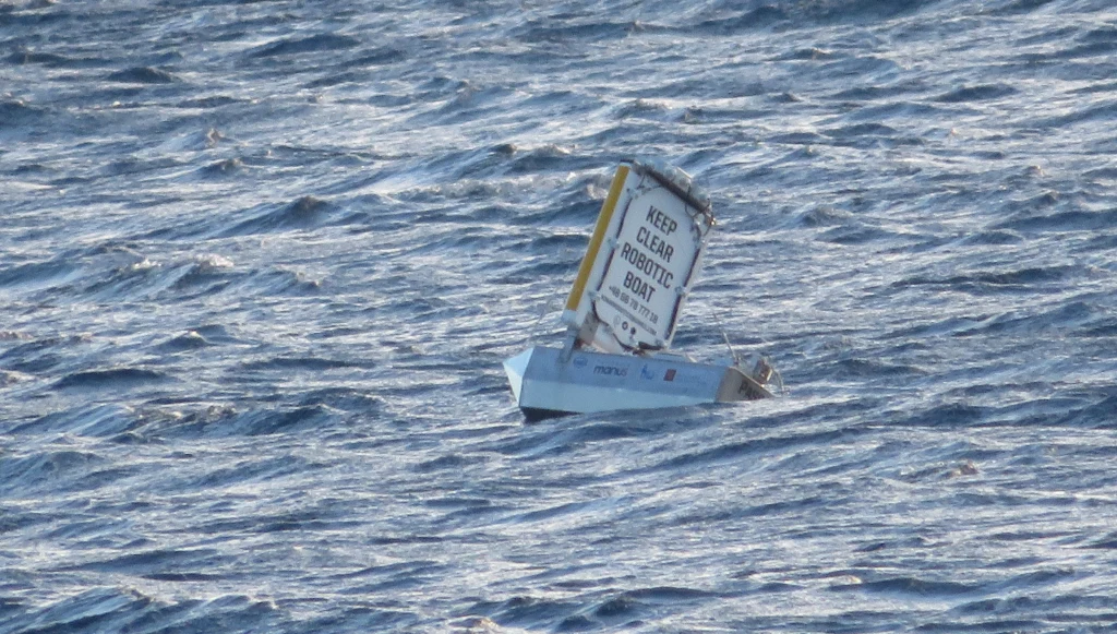 Mała łódź z żaglem pokrytym napisem ostrzegawczym "Keep clear robotic boat" płynie po wzburzonej powierzchni morza. Woda w kolorze ciemnoniebieskim, delikatne fale odbijające światło, wokół brak ludzi.