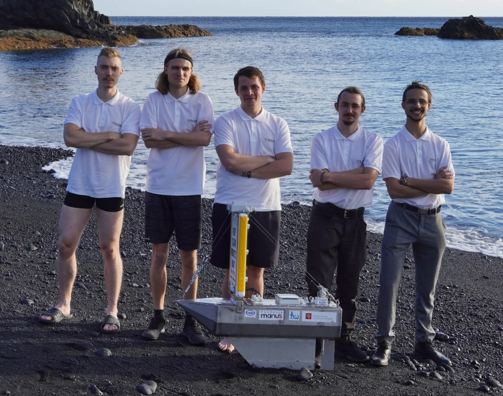 Grupa pięciu młodych mężczyzn ubranych w białe koszule i ciemne spodnie lub krótkie spodenki stoi na czarnej plaży przy brzegu morza, przed nimi znajduje się urządzenie techniczne, przypominające niewielką tratwę lub pojazd badawczy. Na tle spokojnej w...