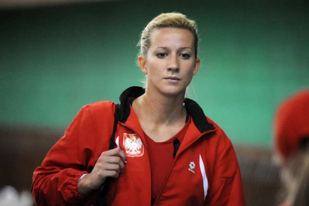 Kobieta w czerwonej sportowej kurtce z orłem na piersi, z blond włosami spiętymi do tyłu, stoi na tle zielonej ściany i skupionym wyrazem twarzy patrzy przed siebie.