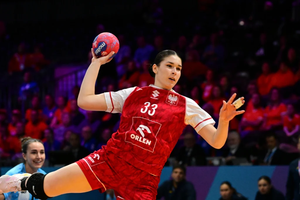 Nikola Głębocka w barwach reprezentacji Polski podczas MŚ w piłce ręcznej Zawodniczka polskiej reprezentacji piłki ręcznej w czerwonym stroju z numerem 33 wyskakuje z piłką do rzutu, na tle rozmazanej publiczności i przeciwniczki w błękitnym stroju.