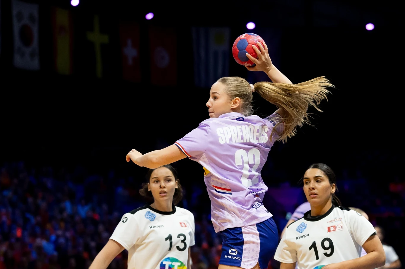 Zoe Sprangers w meczu z Tunezją Zawodniczka w fioletowej koszulce drużyny z numerem 23 wykonuje rzut piłką podczas meczu piłki ręcznej. Obok niej znajdują się dwie przeciwniczki w białych strojach z numerami 13 i 19. W tle widoczne są nieostre flagi narodowe.