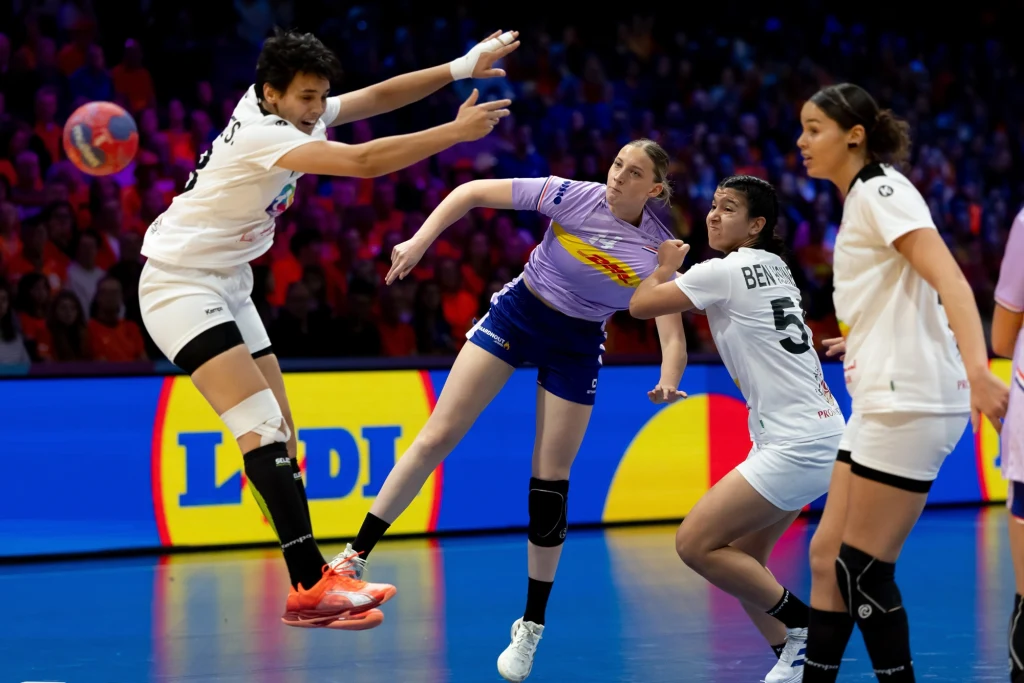 Alieke Van Maurik Zawodniczki drużyn piłki ręcznej walczą o piłkę podczas dynamicznej akcji meczu, w tle widać kolorowe trybuny oraz banery reklamowe.