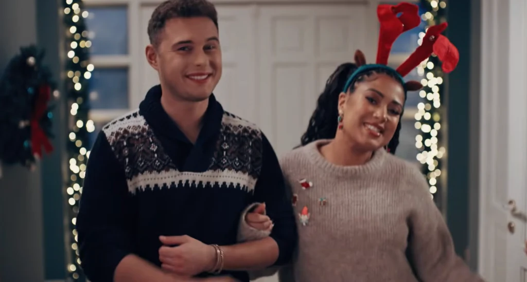 Uśmiechnięta para w świątecznych swetrach stoi razem przy ozdobionych lampkami drzwiach, kobieta ma na głowie opaskę z czerwonymi rogami renifera.