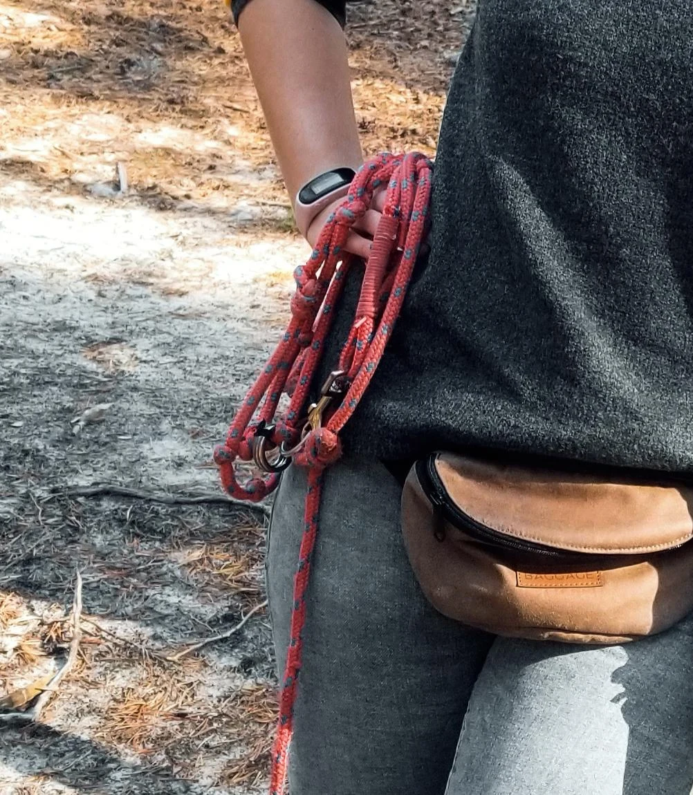 Osoba trzymająca w ręce kolorową linę wspinaczkową, ubrana w ciemny sweter i szare spodnie, z brązową saszetką na biodrach, stojąca na tle leśnego podłoża.