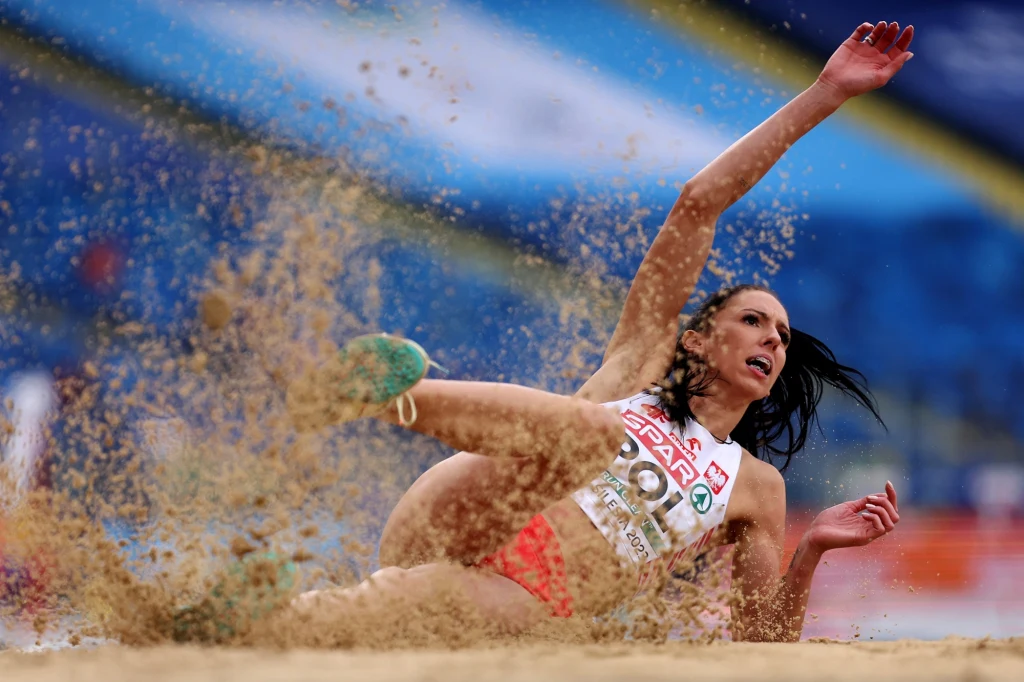 Zawodniczka w trakcie skoku w dal lądująca w piaskownicy, dynamiczne ujęcie momentu kontaktu z piaskiem, widoczne intensywne emocje i ruch, na stroju sportowym numer startowy i polska flaga.