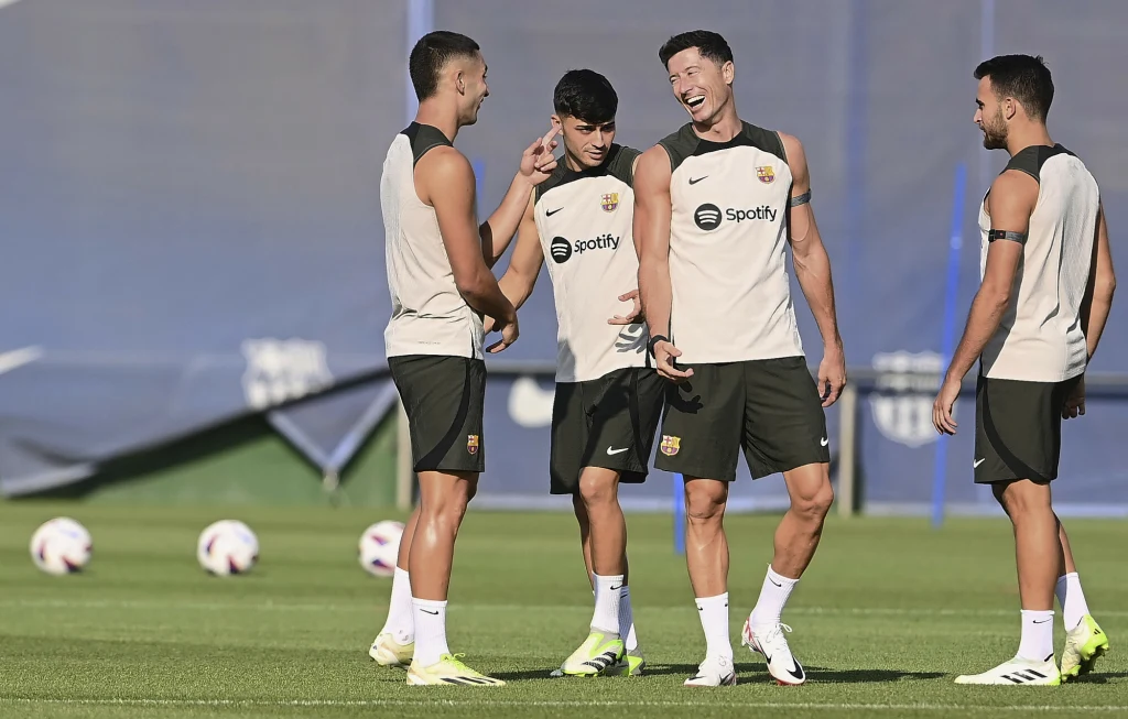 Grupa czterech piłkarzy FC Barcelony uśmiecha się i rozmawia podczas treningu na boisku, ubrani w klubowe stroje treningowe na tle piłek i stadionu.