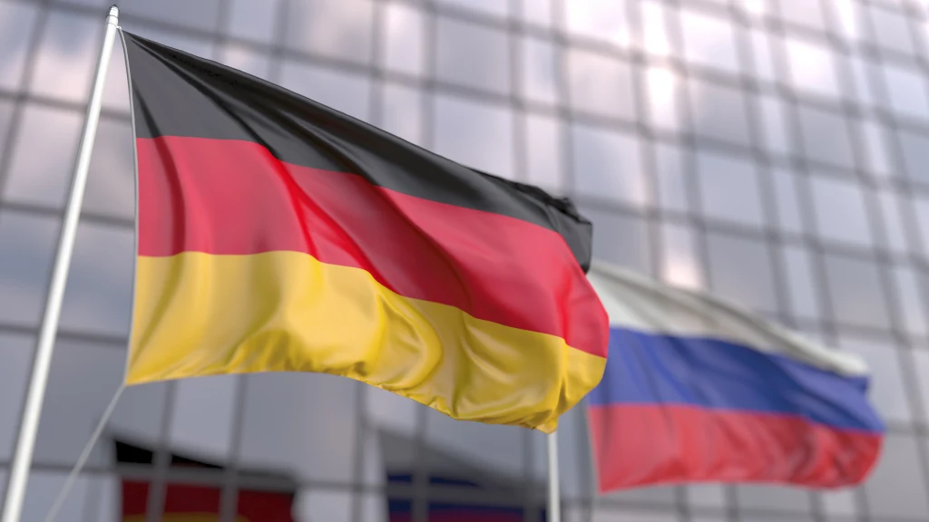 Dwie powiewające flagi: na pierwszym planie niemiecka w barwach czarno-czerwono-żółtych, w tle rosyjska w barwach biało-niebiesko-czerwonych, obie przed nowoczesnym, przeszklonym budynkiem.