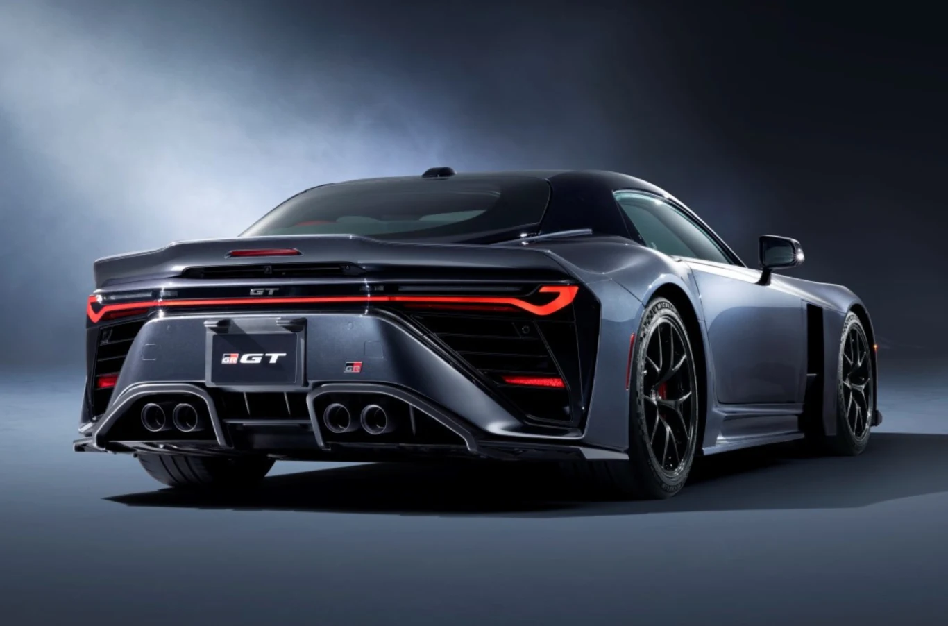 GR GT to pierwsza Toyota z całkowicie aluminiową ramą nadwozia. Ciemnoszary sportowy samochód z widocznym tyłem, światłami LED oraz czterema końcówkami wydechu, stojący na czarnych felgach na tle ciemnego studia.