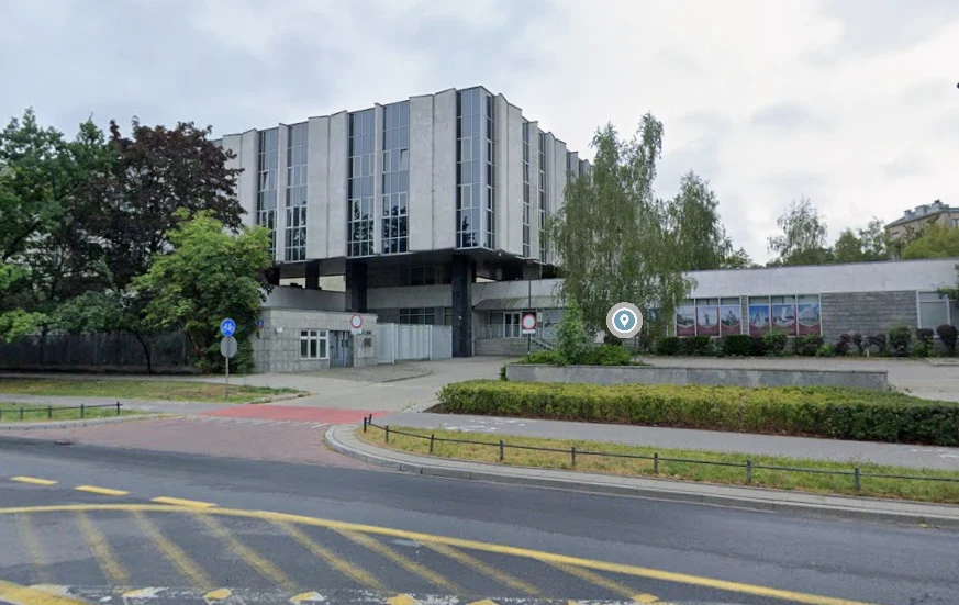 Nowoczesny budynek z przeszkloną fasadą na podwyższonej konstrukcji, otoczony roślinnością i miejską infrastrukturą, w tym chodnikiem i ulicą z oznakowaniem poziomym oraz znakami drogowymi.