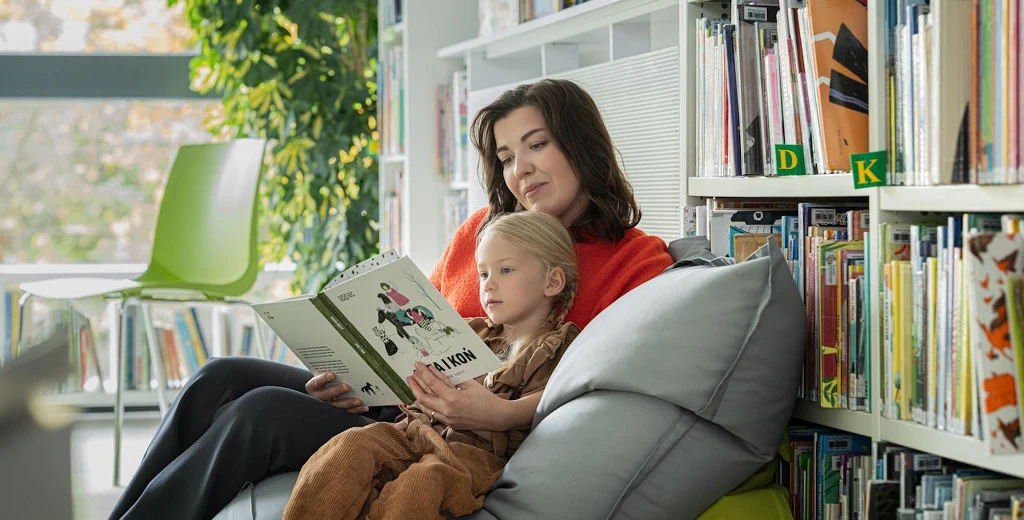 Głośne czytanie dzieciom zwiększa ich zasób słów nawet o 40 proc. Kobieta siedzi z małą dziewczynką na pufie w bibliotece, wspólnie czytają książkę, wokół znajdują się regały z książkami.
