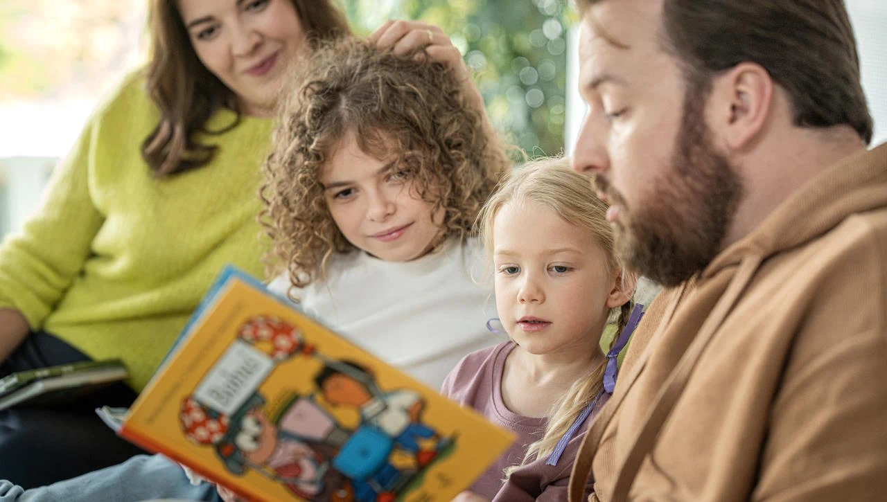 Rodzina spędza wspólnie czas na kanapie, dorosły mężczyzna czyta książkę dwójce dzieci, podczas gdy kobieta z uśmiechem towarzyszy im z boku. Książka ma kolorową okładkę z ilustracjami.