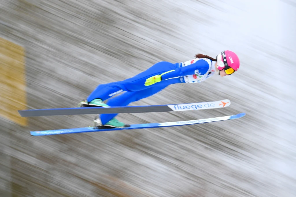 Skoczek narciarski ubrany w niebieski kombinezon i różowy kask podczas lotu na tle rozmytego śniegu, sylwetka wyciągnięta w aerodynamicznej pozycji.