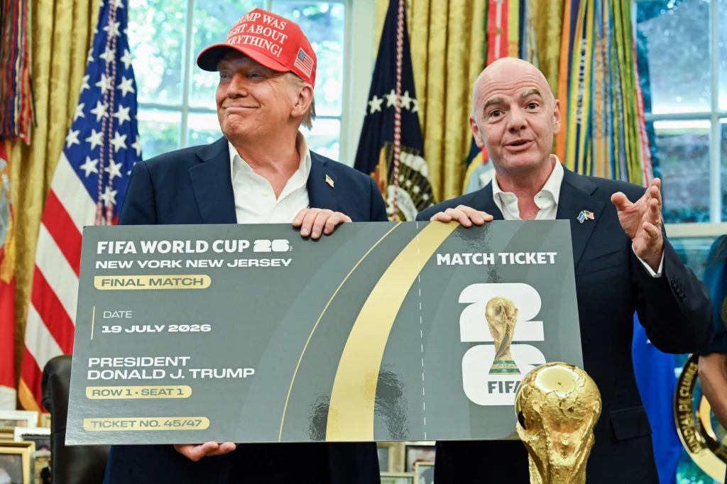 Donald Trump i Gianni Infantino Dwóch mężczyzn trzyma dużą symboliczną wejściówkę na finał Mistrzostw Świata FIFA 2026 w Nowym Jorku, za nimi znajdują się amerykańskie flagi oraz złota statuetka Pucharu Świata. Jeden z nich ma czerwoną czapkę z napisem.