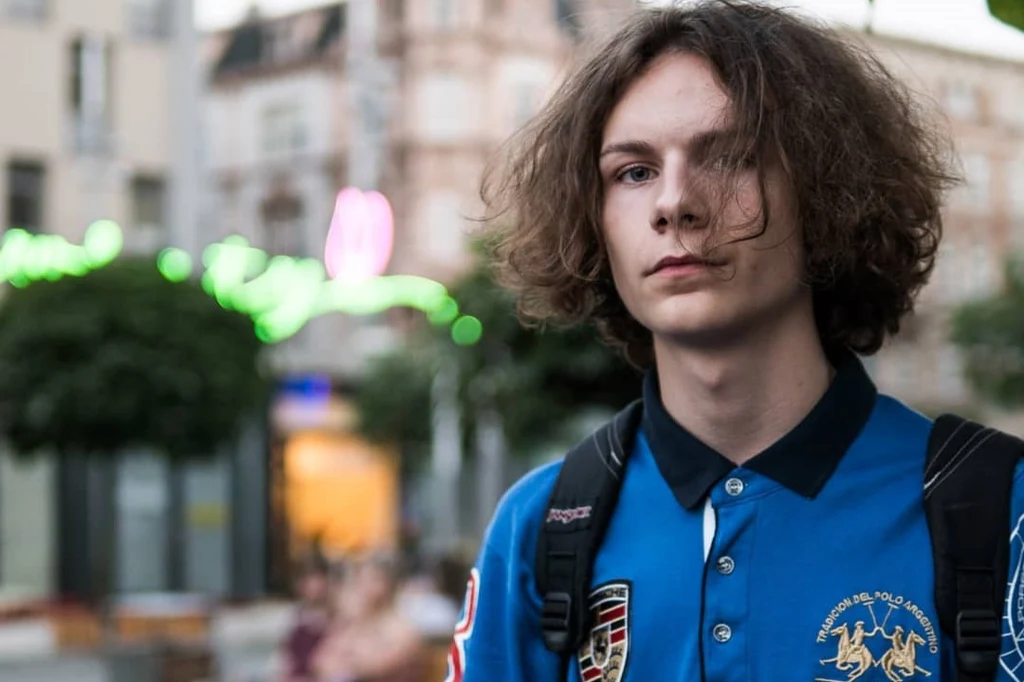 Schafter wydał album "Palimpsest" Młody mężczyzna z długimi, kręconymi włosami w niebieskiej koszulce polo z naszywkami i czarnym plecakiem na ramionach, stoi na tle rozmytego miasta o zmierzchu.