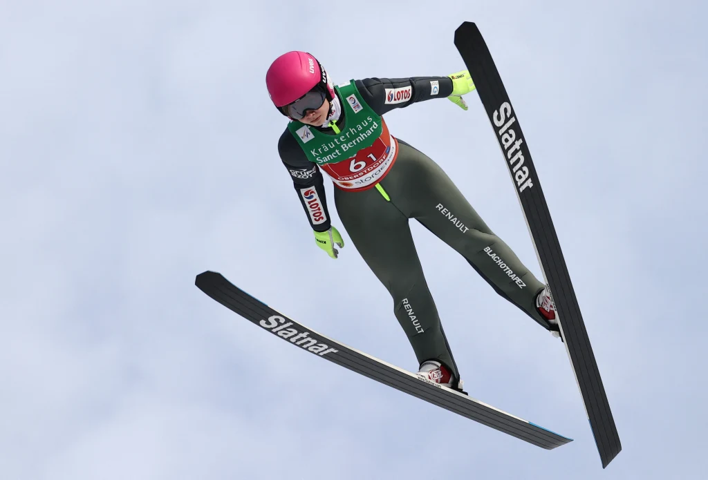 Zawodnik w locie podczas skoku narciarskiego na tle nieba, ubrany w kombinezon sportowy i różowy kask, trzyma równowagę z wyciągniętymi nartami.