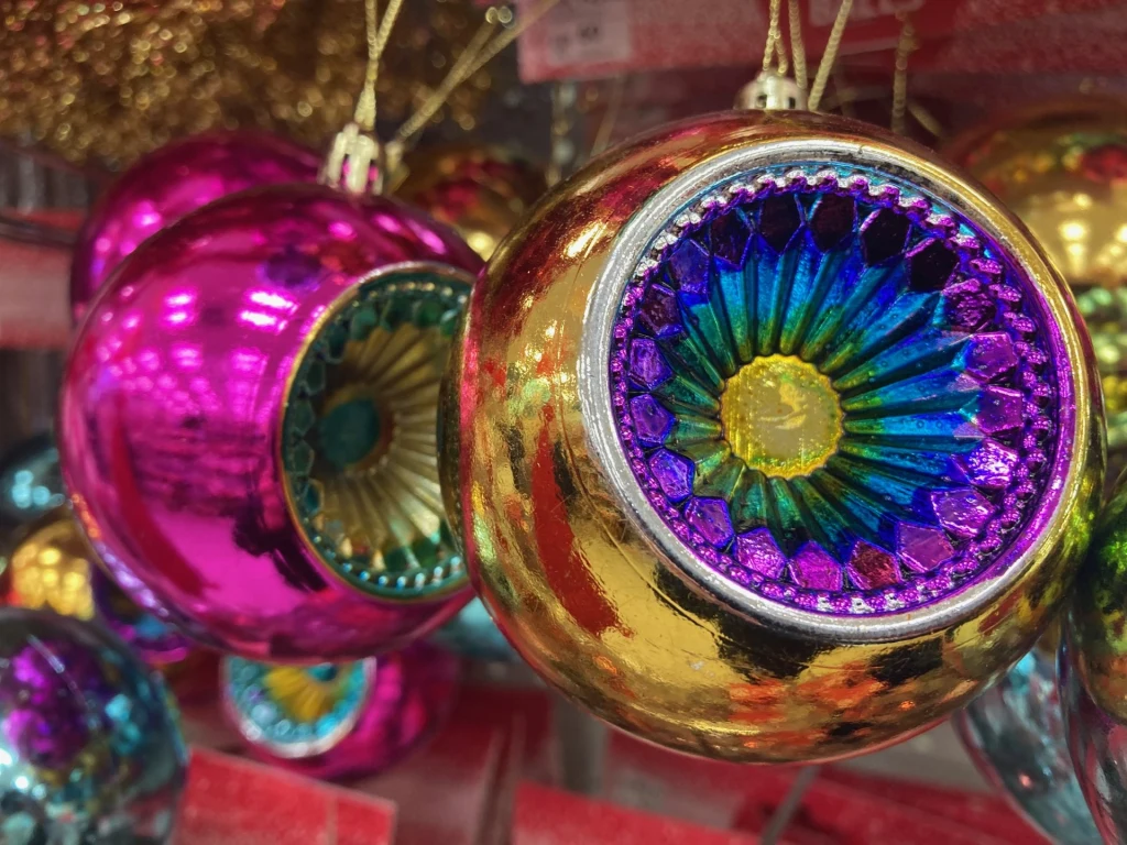 Te bombki mogą być doskonałym kolorystycznym akcentem na stonowanej choince Kolorowe bombki choinkowe o błyszczących powierzchniach w odcieniach złota, fioletu, różu i niebieskiego, z wypukłymi środkami w kształcie kwiatu, zawieszone na złotych sznurkach.