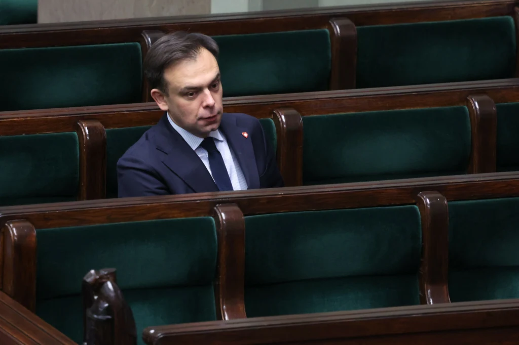 Mężczyzna w garniturze siedzący samotnie w zielonych ławach sejmowych, pogrążony w zadumie.