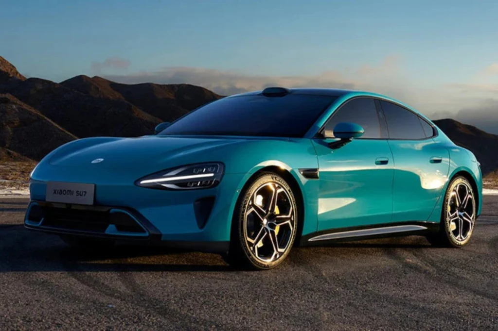 Turkusowy, nowoczesny samochód elektryczny zaparkowany na tle gór o zachodzie słońca. Reflektory LED, sportowe felgi oraz aerodynamiczna sylwetka podkreślają futurystyczny charakter auta. Karoseria błyszczy w delikatnym świetle, odbijając otoczenie.