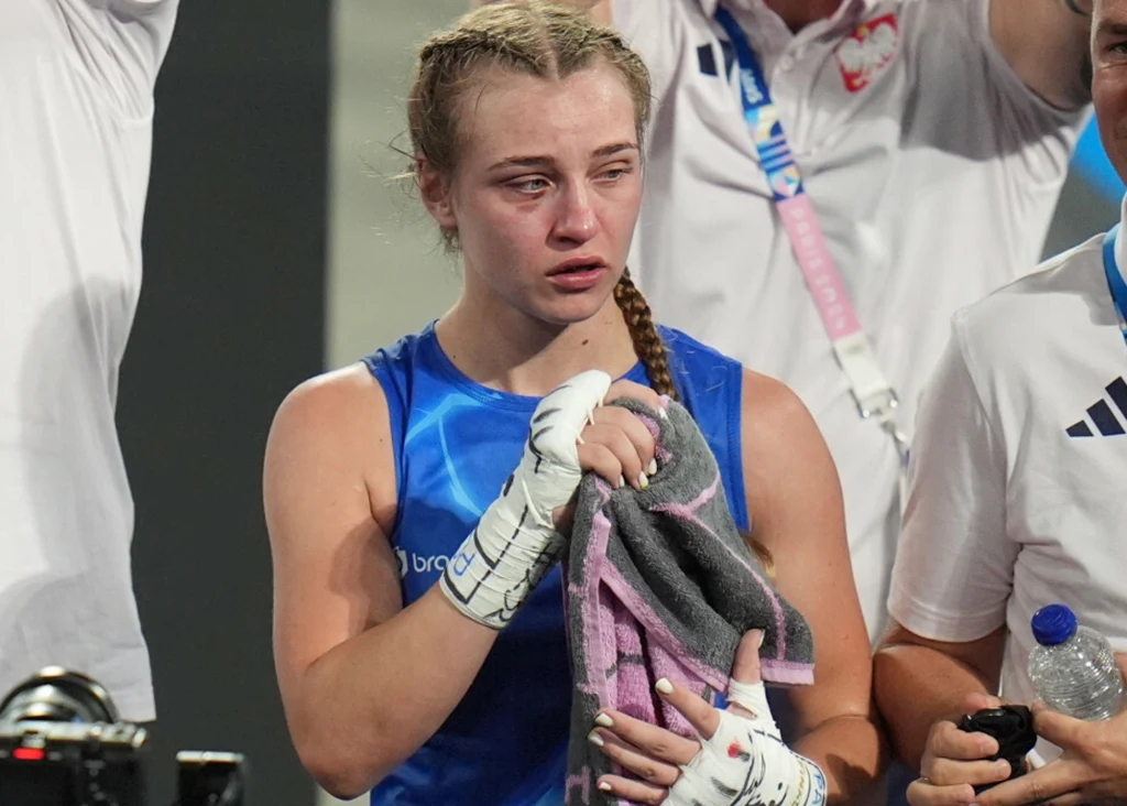 Zmęczona młoda kobieta w sportowym stroju bokserskim oraz bandażami na dłoniach trzyma ręcznik, na jej twarzy widoczna jest silna emocja i łzy, a w tle stoją członkowie ekipy w białych koszulkach.
