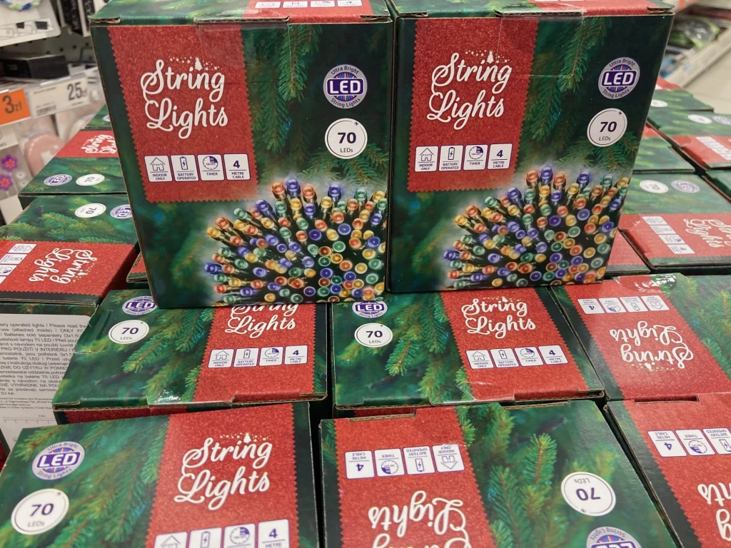 Kolorowe lampki na choinkę właśnie powróciły do sklepów Kolorowe opakowania lampek świątecznych LED ułożone w stos, dominują motywy zieleni oraz czerwieni, na opakowaniach widoczne informacje o liczbie lampek i ich właściwościach.