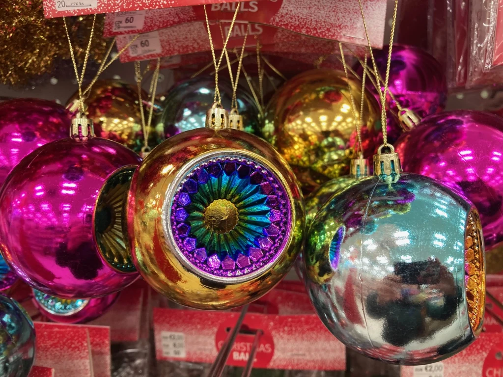 Urocze bombki stylizowane na lata 90. będą świetnym dodatkiem do choinki w klasycznym wydaniu Kolorowe bombki choinkowe w różnych odcieniach różu, złota, srebra i zieleni zawieszone na sznurku, w tle półka sklepu z oznaczeniami cenowymi. Jedna z bombek posiada dekoracyjny wzór przypominający oko.