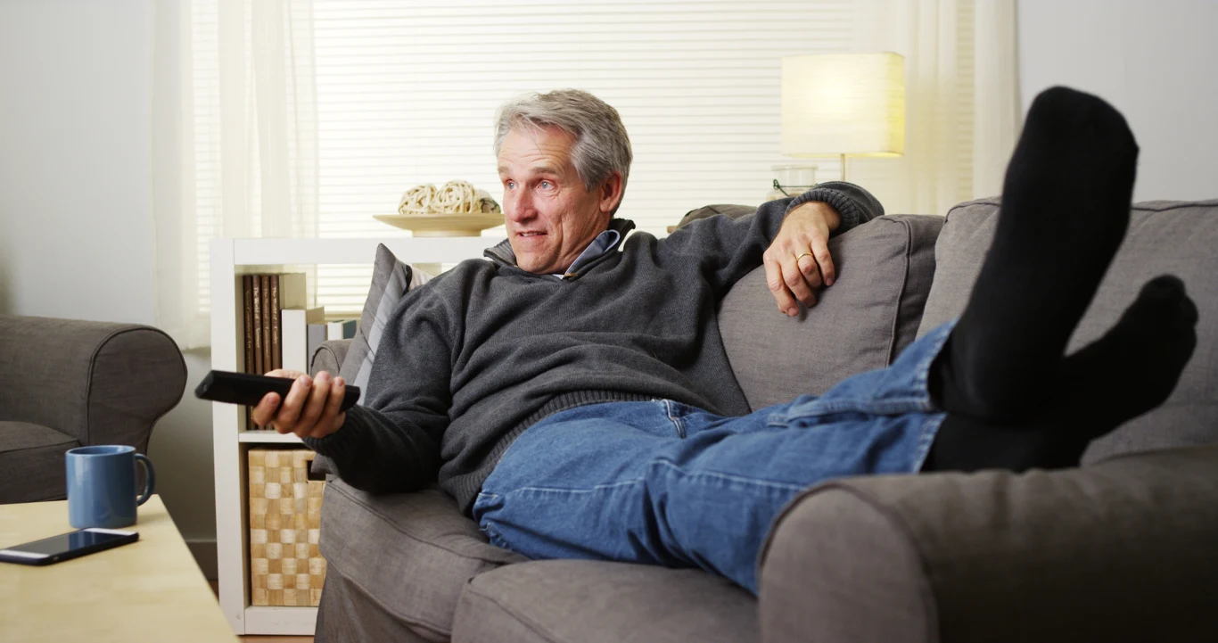 Starszy mężczyzna w luźnym swetrze i dżinsach wygodnie leży na kanapie z pilotem w ręku, patrząc w telewizor. W tle jasne wnętrze salonu z lampą i kubkiem na stole.