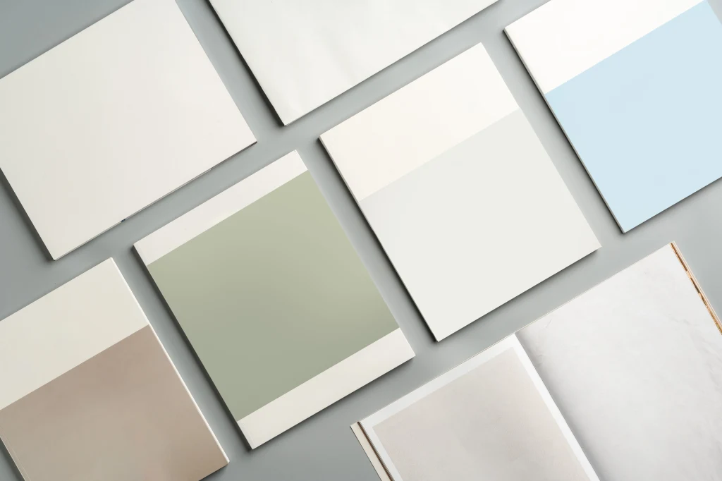 Purdowe pastele i zgaszone kolory będą towarzyszyć "Tańczącemu w chmurach" Równomiernie rozłożone próbki kolorów, głównie w pastelowych i jasnych odcieniach, rozmieszczone na szarej powierzchni. Po prawej stronie widoczna jest otwarta, pusta książka lub katalog.