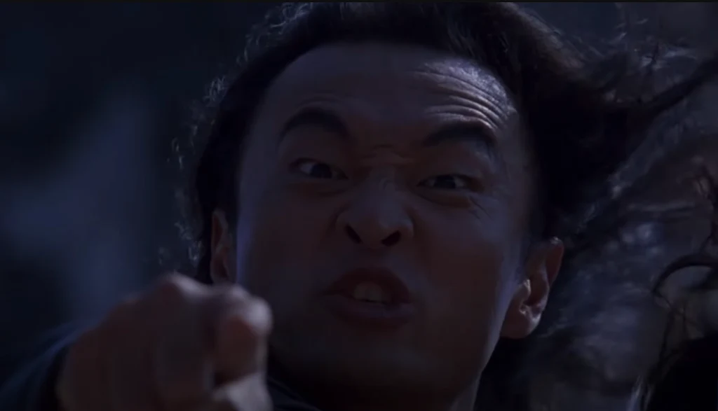 Twarz mężczyzny z wyraźnie zaciśniętymi brwiami, szeroko otwartymi oczami i ustami wyrażającymi krzyk lub gniew. Wskazuje palcem w stronę obiektywu, włosy rozwiane przez wiatr, ciemne tło potęguje dramatyzm sceny.