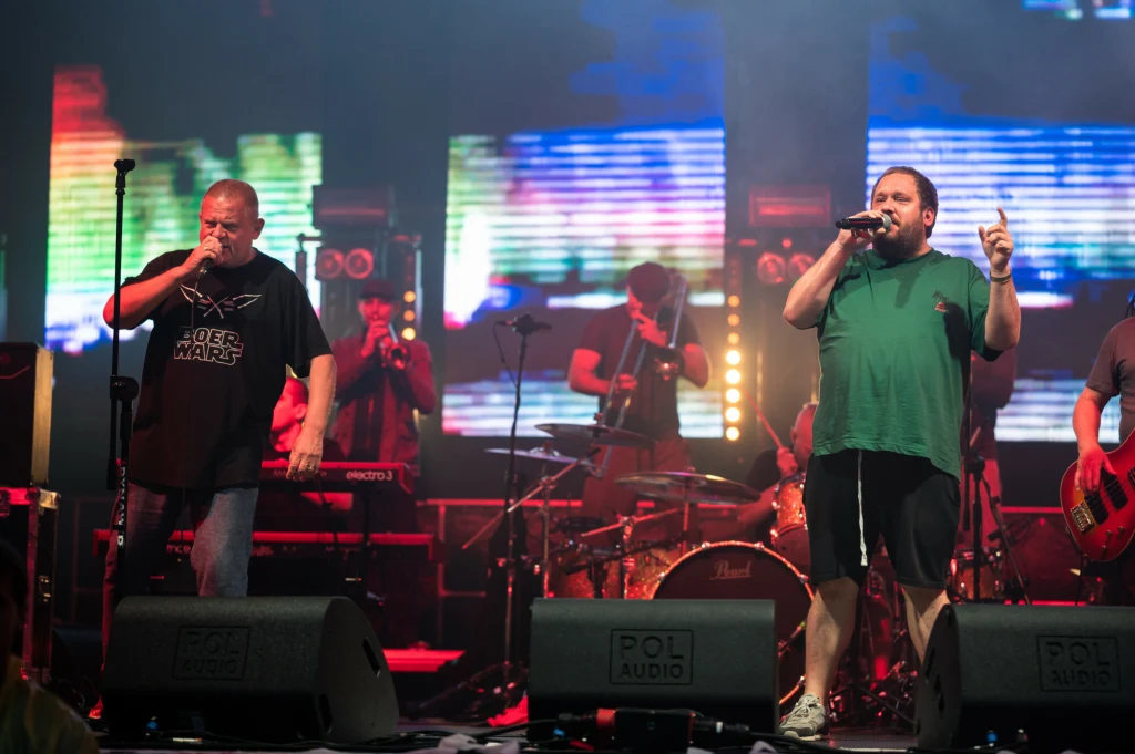 Zespół muzyczny występujący na scenie, dwóch wokalistów z mikrofonami na pierwszym planie, za nimi muzycy grający na gitarze, perkusji i instrumentach dętych, kolorowe światła sceniczne i ekrany LED w tle.