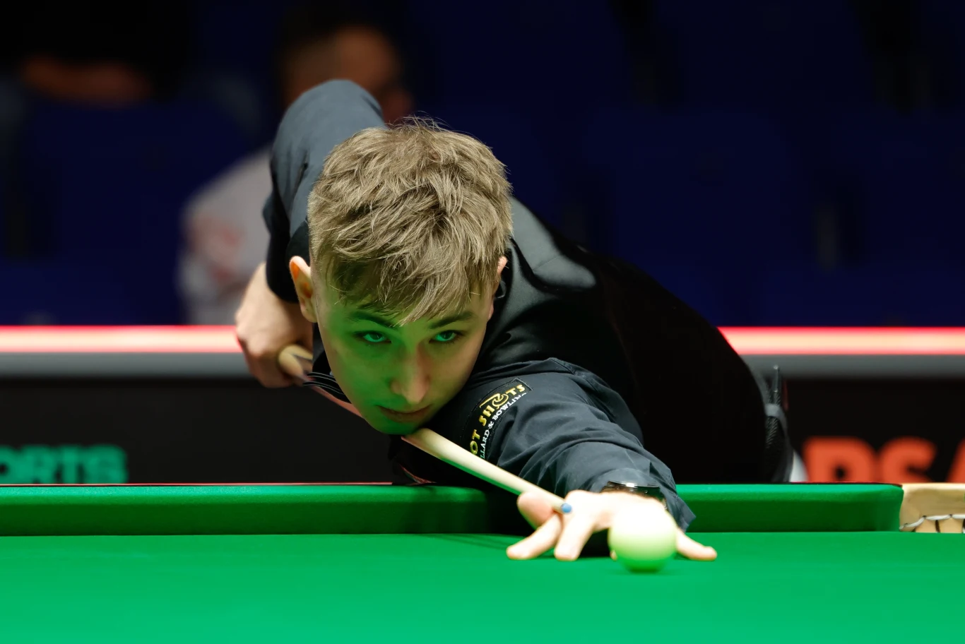 Antoni Kowalski Młody mężczyzna skupiony na wykonaniu precyzyjnego uderzenia podczas meczu snookera, trzyma kij bilardowy i celuje w białą bilę na zielonym stole; w tle niewyraźni widzowie.