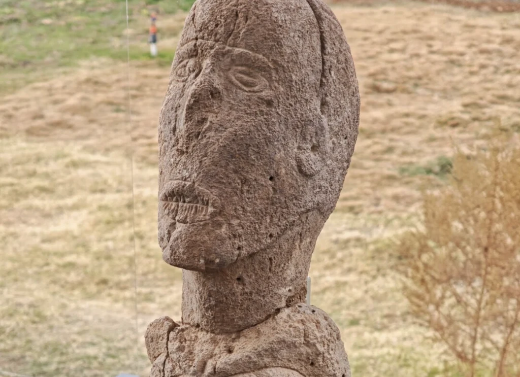 Tajemnicza figurka z zaszytymi ustami sprzed 11 000 lat. Turcja odkrywa kolejne tajemnice Stara kamienna rzeźba przedstawiająca ludzką postać z wyraźnie zarysowanymi rysami twarzy, ustawiona na tle trawiastego terenu.