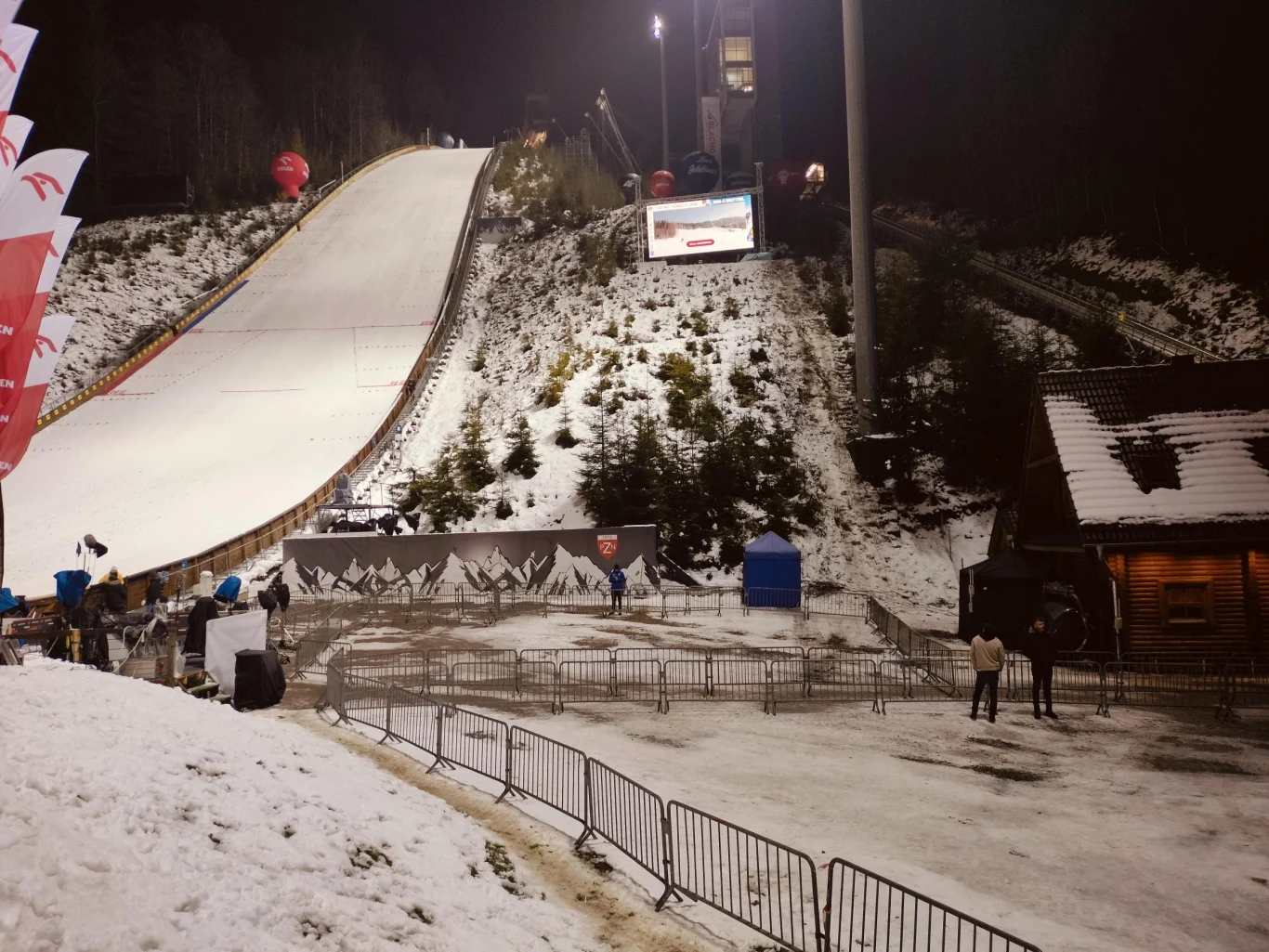 Pustki na skoczni w Wiśle w czasie konkursu kobiet Duża skocznia narciarska oświetlona sztucznym światłem, pokryta śniegiem, otoczona lasem i trybunami. Widać barierki zabezpieczające, niewielką drewnianą budowlę i niewielką grupę osób przygotowującą się do wydarzenia.