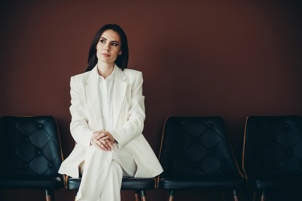 W modzie biel jest jednym z kolorów bazowych Kobieta w białym garniturze siedzi na krześle na tle ciemnej ściany, obok niej puste czarne krzesła, jej wyraz twarzy jest spokojny i skoncentrowany.