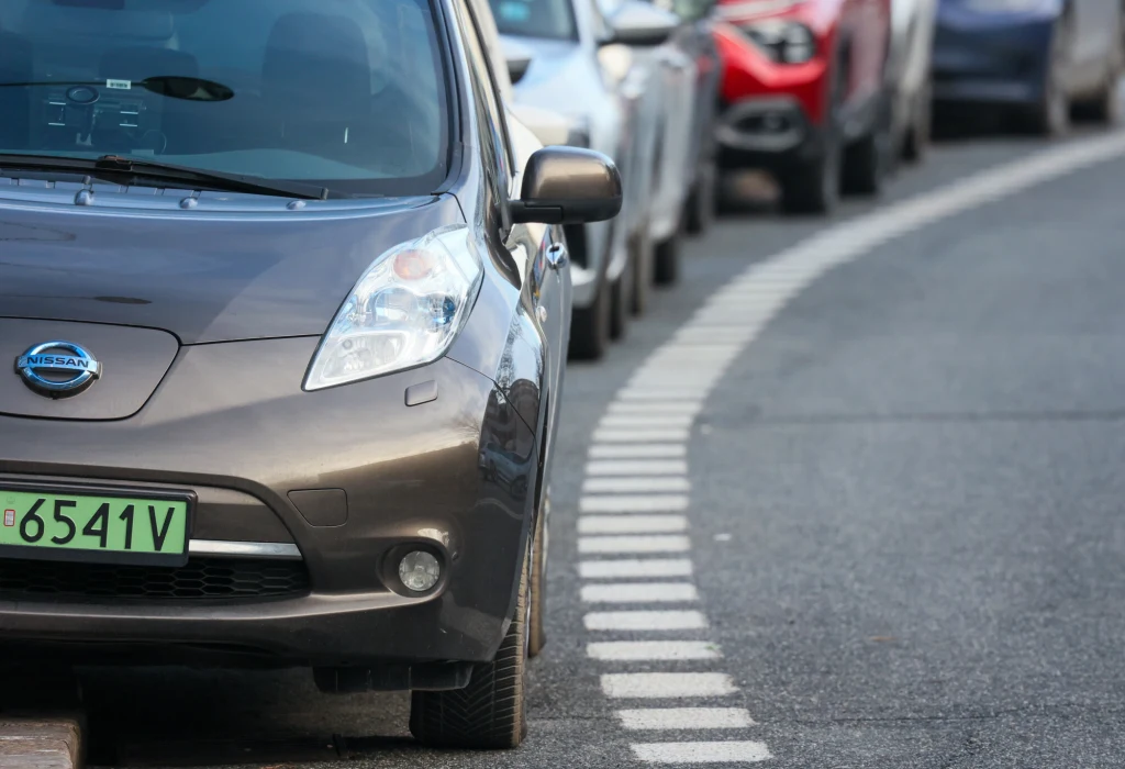 Szereg zaparkowanych samochodów ustawionych wzdłuż ulicy, na pierwszym planie widoczny przód szarego samochodu marki Nissan, z zieloną tablicą rejestracyjną.