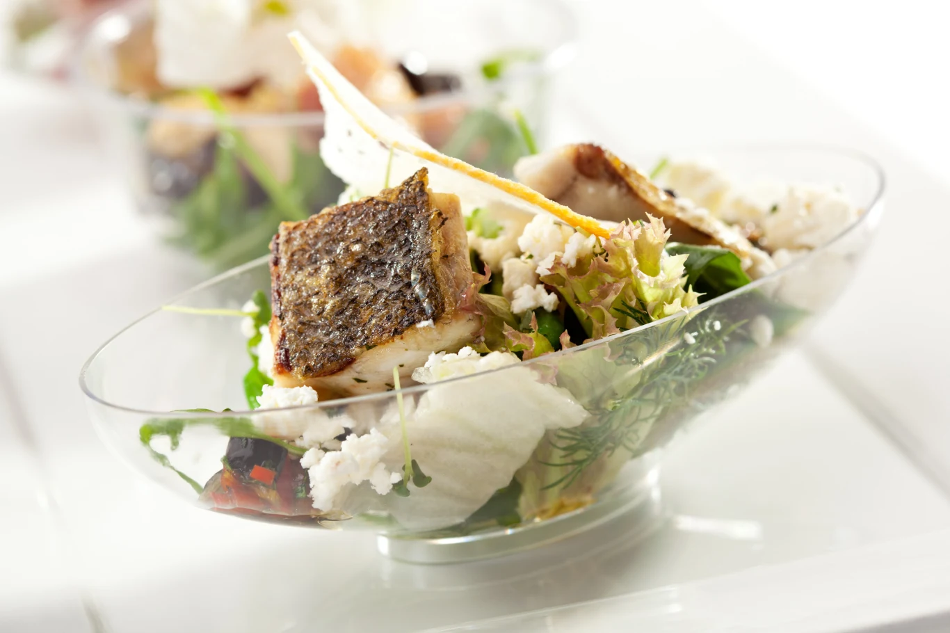 Kawałki smażonej ryby z chrupiącą skórką, świeże liście sałaty, pokruszony biały ser, zioła i chrupiące dodatki podane w przezroczystej miseczce.