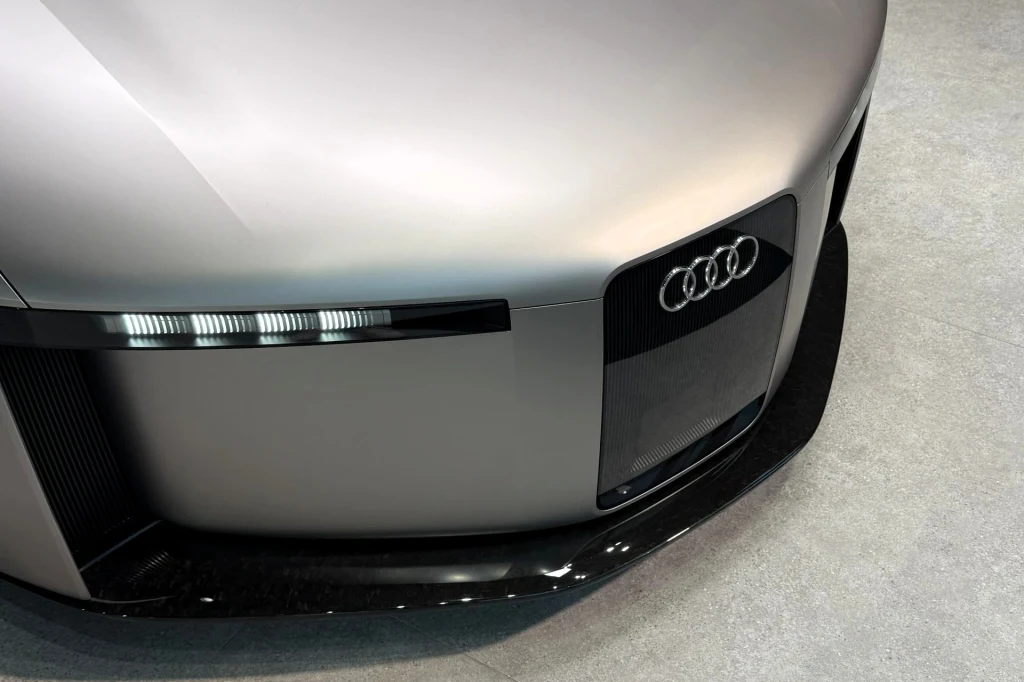 Przód futurystycznego samochodu Audi z charakterystycznym logo, nowoczesnym czarnym grillem i wąskimi listwami świateł LED na tle jednolitej, jasnej podłogi.