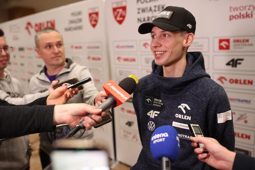 Kacper Tomasiak Młody sportowiec w sportowej bluzie i czapeczce udziela wywiadu przedstawicielom mediów, wokół niego kilka mikrofonów z logotypami różnych stacji. W tle plansze z logotypami sponsorów, a obok niego kilka osób, w tym dziennikarze.