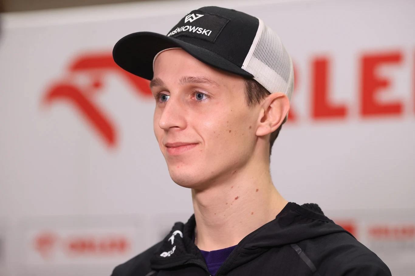 Paweł Wąsek Młody mężczyzna w czapce z daszkiem i sportowej kurtce, stoi na tle rozmazanego logotypu wyglądającego na sportowy lub promocyjny. Uśmiecha się lekko, patrząc nieco w bok.