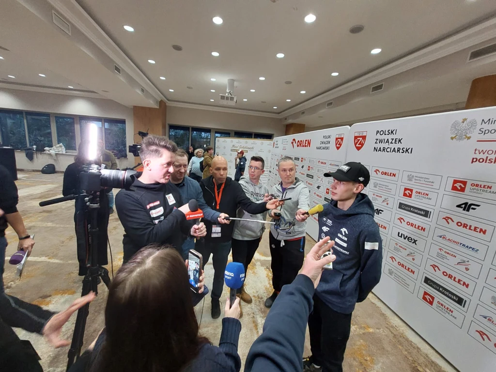 Kacper Tomasiak budzi zainteresowanie mediów Kilka osób udziela wywiadu dziennikarzom przed ścianką z logotypami Polskiego Związku Narciarskiego oraz sponsorów, wokół rozmówcy zgromadzona jest grupa reporterów z mikrofonami i kamerami, całość odbywa się w oświetlonej sali konferencyjnej.