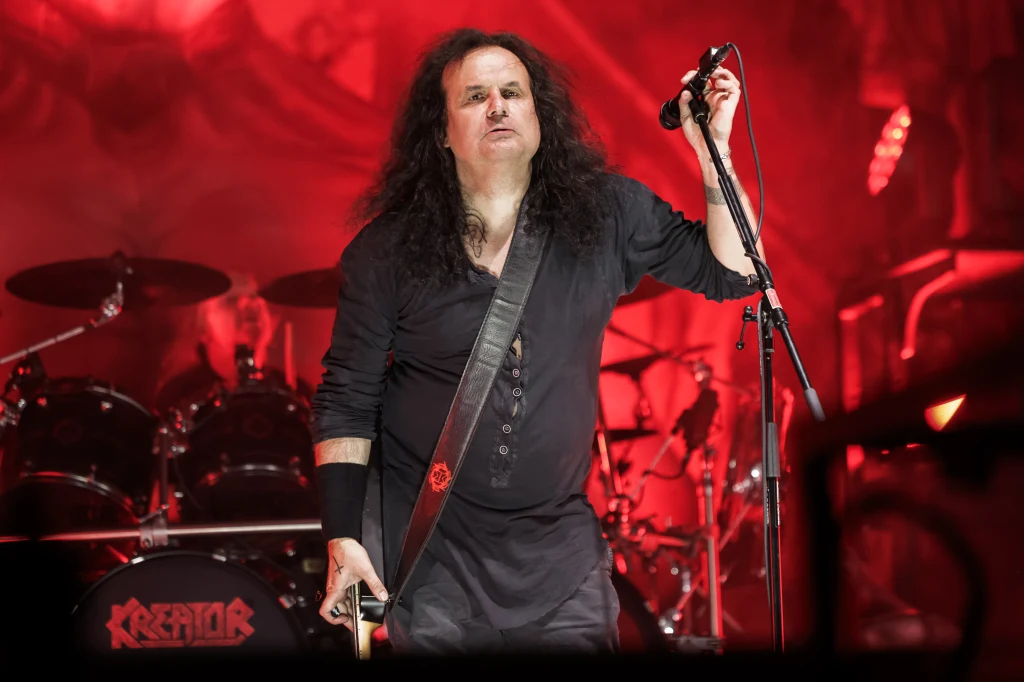 Mille Petrozza (Kreator) w akcji Wokalista zespołu metalowego stoi na scenie z mikrofonem w dłoni, otoczony przez czerwone światła sceniczne i perkusję w tle, na której widoczna jest nazwa zespołu Kreator.