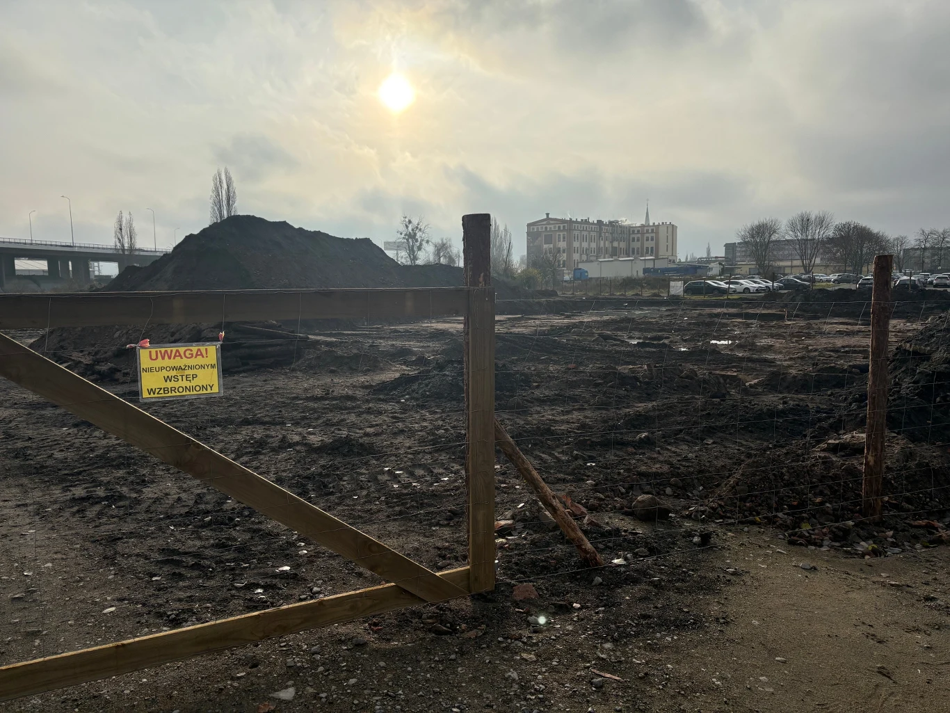 Ograniczony teren budowy ogrodzony drewnianą furtką i metalową siatką, ziemia rozkopana, widoczne hałdy ziemi oraz żółta tabliczka ostrzegawcza przy wejściu, w tle zabudowania miejskie, na niebie zachmurzenie i przebijające się słońce.