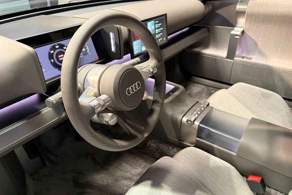 Wnętrze nowoczesnego samochodu Audi z wyraźnie widoczną kierownicą, cyfrowymi wyświetlaczami na desce rozdzielczej oraz eleganckimi, jasnymi fotelami i minimalizmem wykończenia kokpitu.