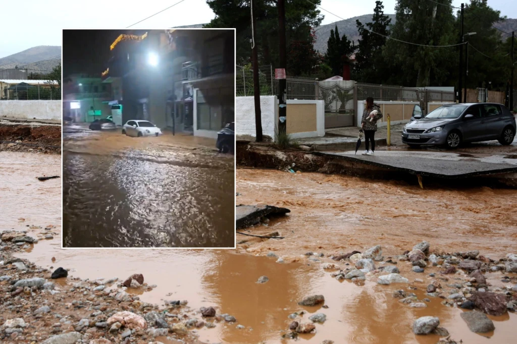 Ulice zalane brunatną wodą po gwałtownej powodzi, zniszczone fragmenty nawierzchni, samochód częściowo unieruchomiony przez wodę, dwie osoby obserwujące sytuację z chodnika, w tle budynki mieszkalne i samochód osobowy.