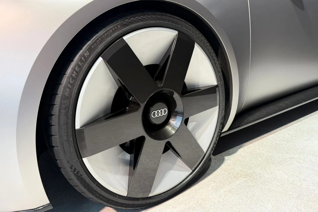 Duże koło samochodu marki Audi z charakterystycznym logo pośrodku, felga w nowoczesnym, geometrycznym stylu z szerokimi czarnymi ramionami, widoczna część nadwozia w kolorze srebrnym.