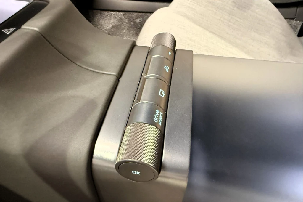 Panel sterowania z przyciskami i pokrętłem zamontowany na konsoli samochodu. Widoczne napisy oraz symbole dotyczą funkcji drzwi i okien pojazdu.