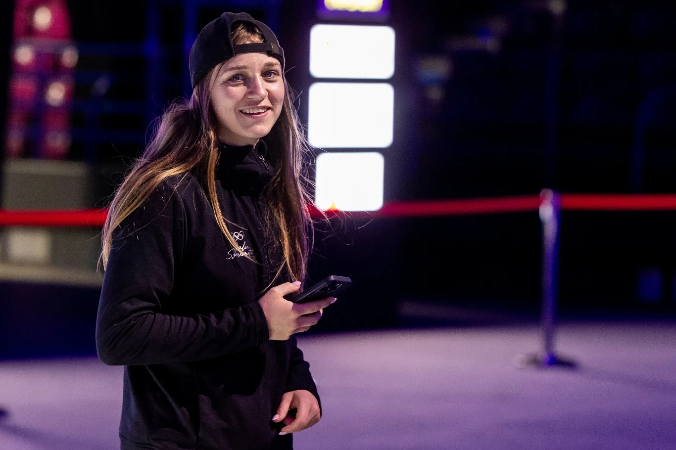 Julia Szeremeta Młoda kobieta w sportowej bluzie i czapce z daszkiem, trzymająca w ręku telefon, uśmiechnięta, stoi w lekko oświetlonym wnętrzu z barierkami i neonowymi światłami w tle.